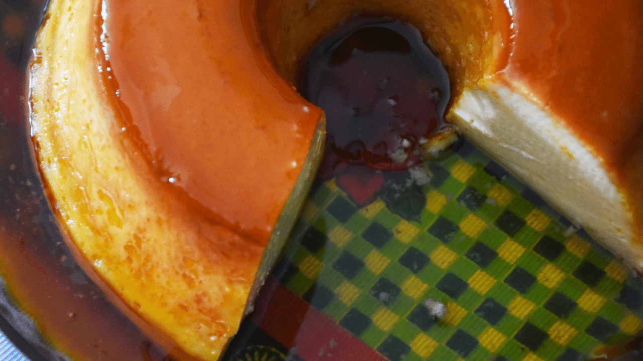 Pudim de padaria feito e aparecendo de cima cortado e banhado na calda de caramelo num travessa de pudim transparente em cima de guardanapo xadrez com cores preto, amarelo e verde.