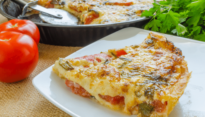 torta de liquidificador servido num prato branco quadrado com duas frutas de tomates aparecendo a esquerda e a metade da forma aparecendo no fundo